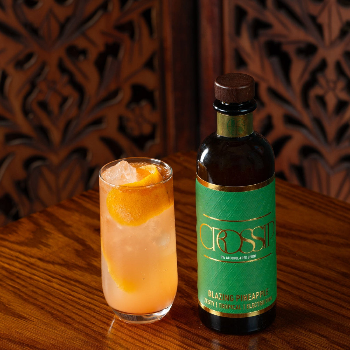 A bottle of Crossip Blazing Pineapple cocktail syrup stands next to a prepared cocktail in a tall glass on a wooden table, with a carved wooden background.