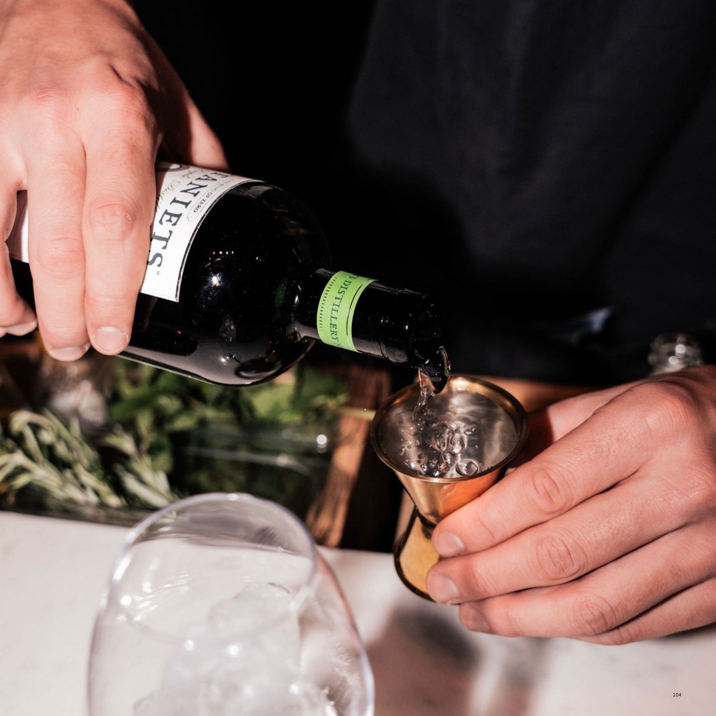 A close-up lifestyle shot of a bartender pouring a dark liquid from a bottle labeled 'BOTANIETS' into a copper jigger, with ice in a glass in the foreground.