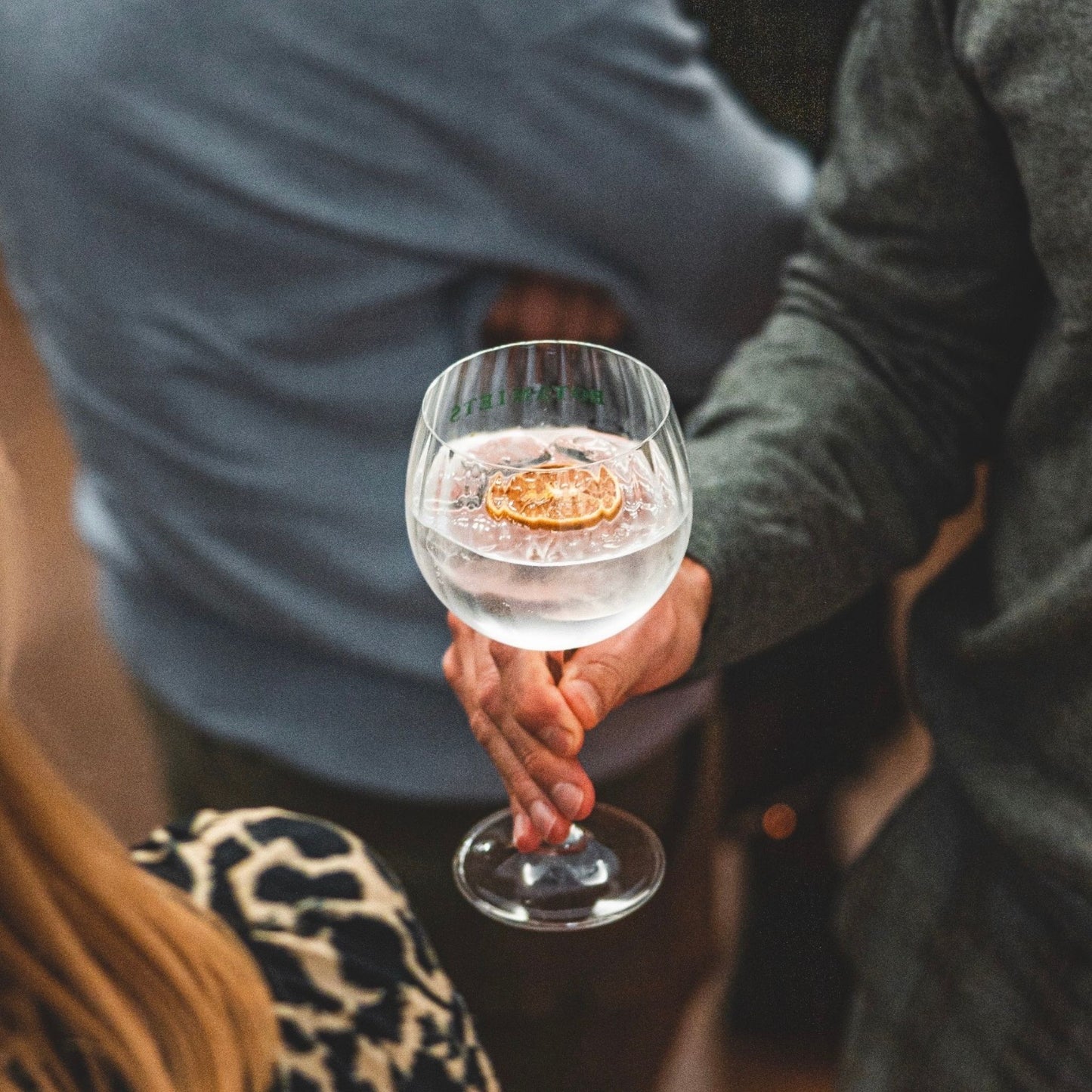 A clear stemmed wine glass with a dried orange slice floating in a clear drink, held by a person in a dark gray sweater at a social gathering.