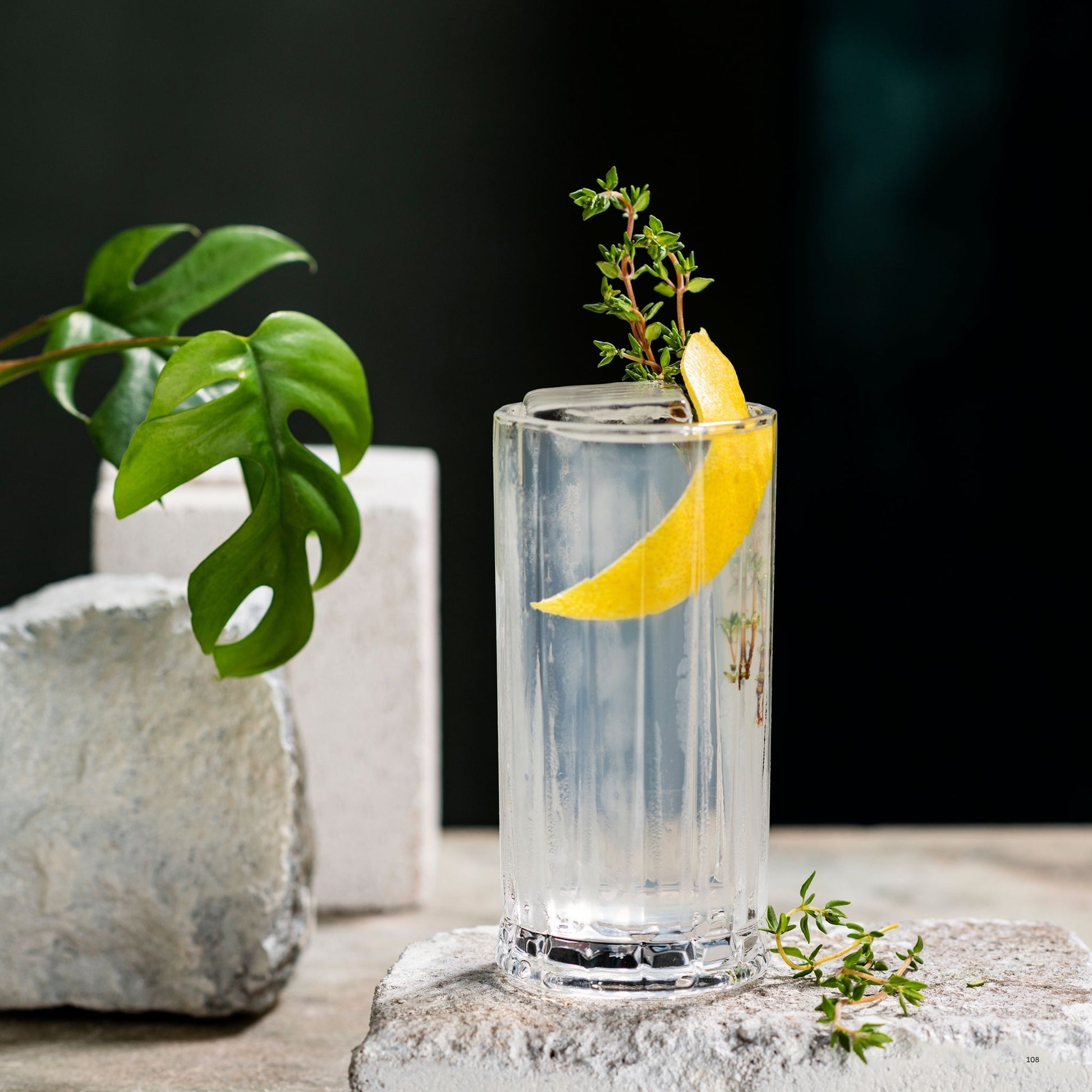 A clear highball glass filled with a transparent drink, garnished with a yellow lemon slice and a small herb sprig, styled on a stone surface with green leaves and white blocks in the background.
