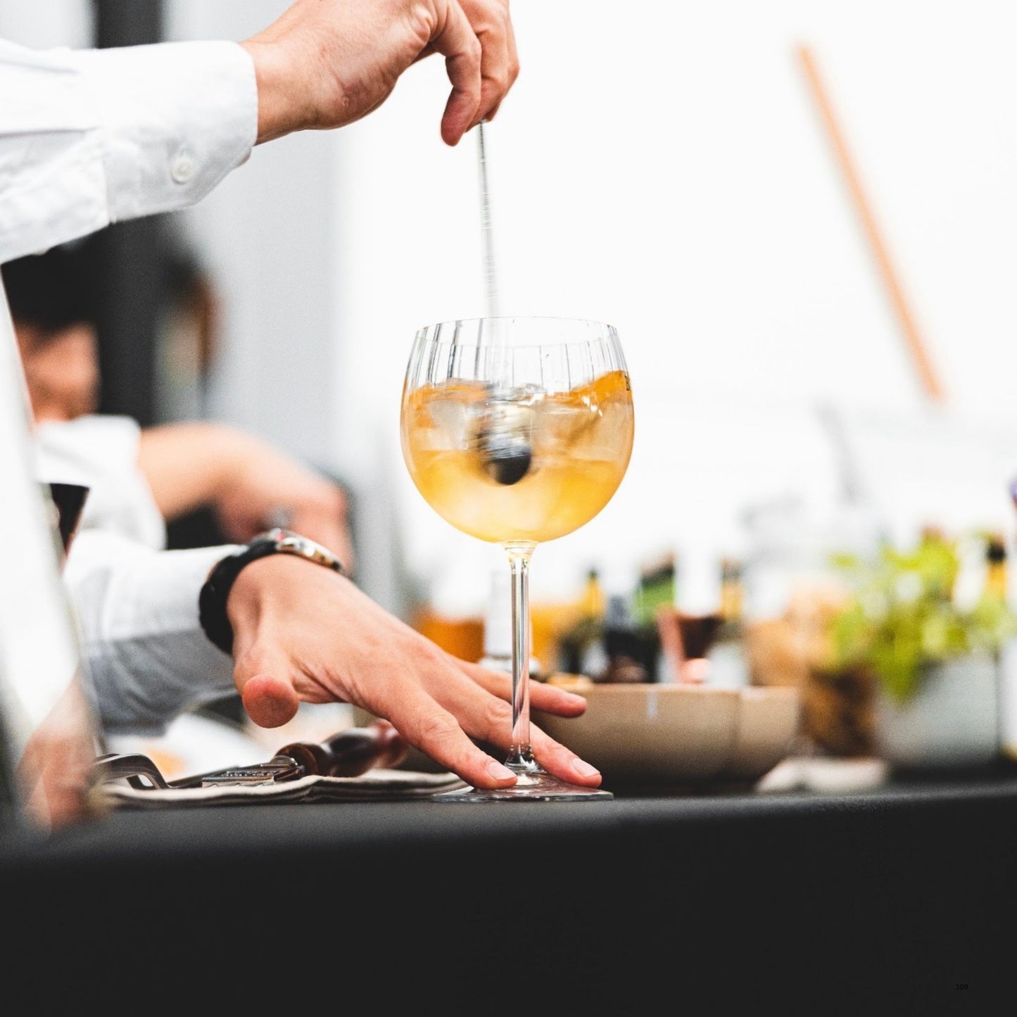 A clear stemmed cocktail glass containing an amber drink with ice, held by a bartender whose hands and white shirt are visible, with a softly blurred bar background.