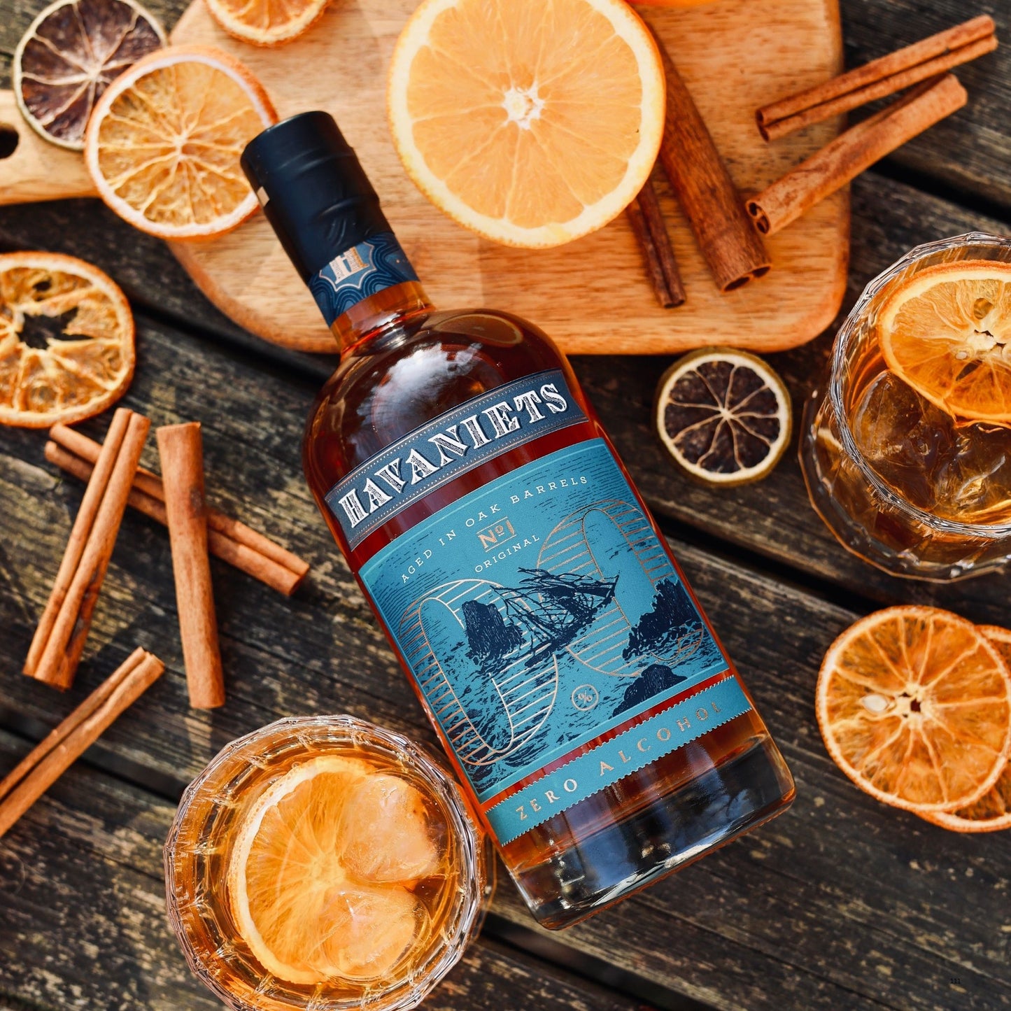 A bottle of Havaniets Zero Alcohol rum spirit is arranged on a rustic dark wooden table with dried orange slices, cinnamon sticks, and glasses of the drink.