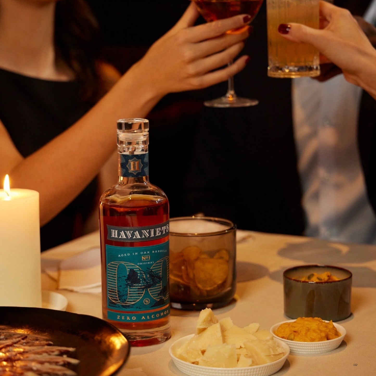 A bottle of Havaniets Zero Alcohol mixed drink mix is displayed on a table with snacks, chips, and a lit candle, while people in the background raise glasses in a celebratory toast.
