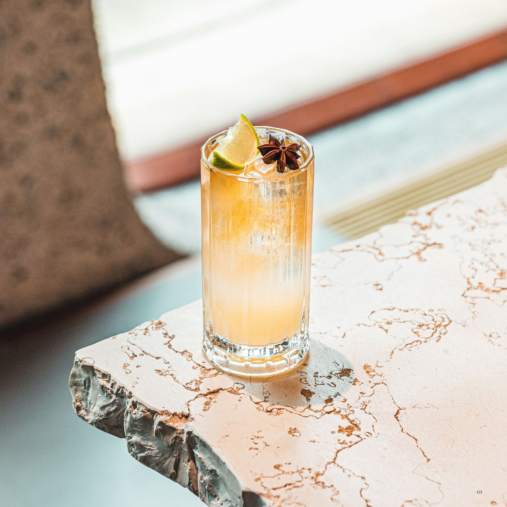 A clear highball glass filled with a pale golden beverage, garnished with a lime wedge and a dark star anise, set on a distressed white table in a softly blurred indoor setting.