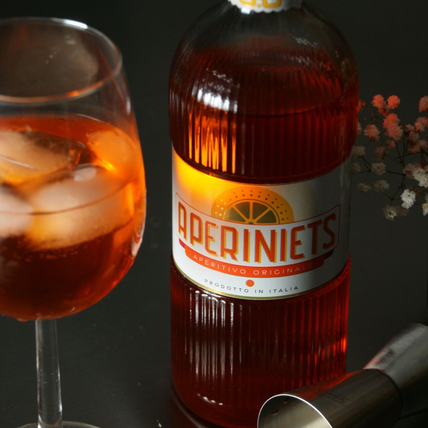 A bottle of Aperiniets Aperitivo Original non-alcoholic Italian drink with a glass of the same amber-red liquid and ice, styled on a dark surface with a jigger in the foreground.