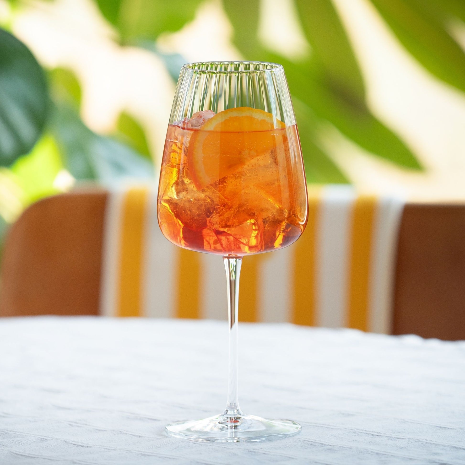 A clear stemmed wine glass with a flared rim, filled with an orange cocktail and a lemon slice, set on a white tablecloth with a blurred outdoor patio background.