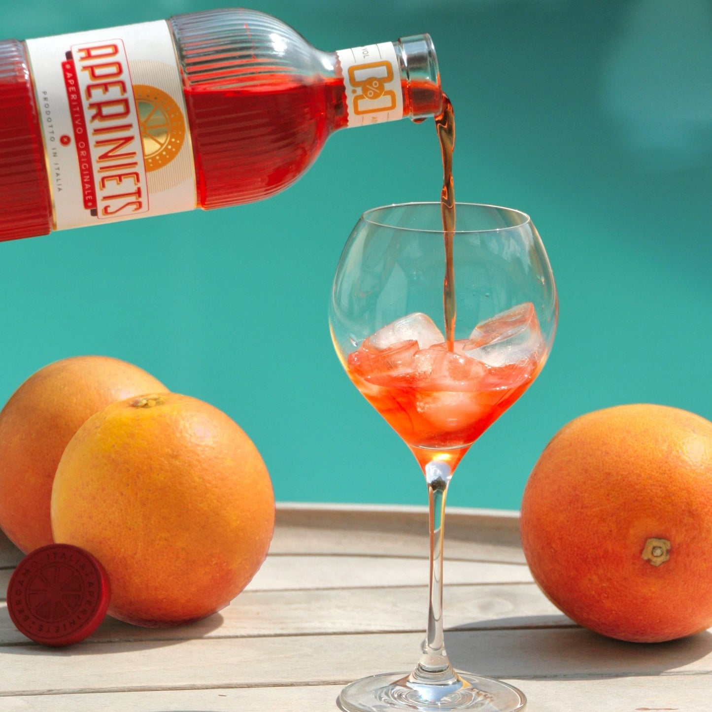 A bottle of Apernièts red liqueur is pouring its dark red liquid into a stemmed glass filled with ice, set on a wooden surface with grapefruits against a teal background.