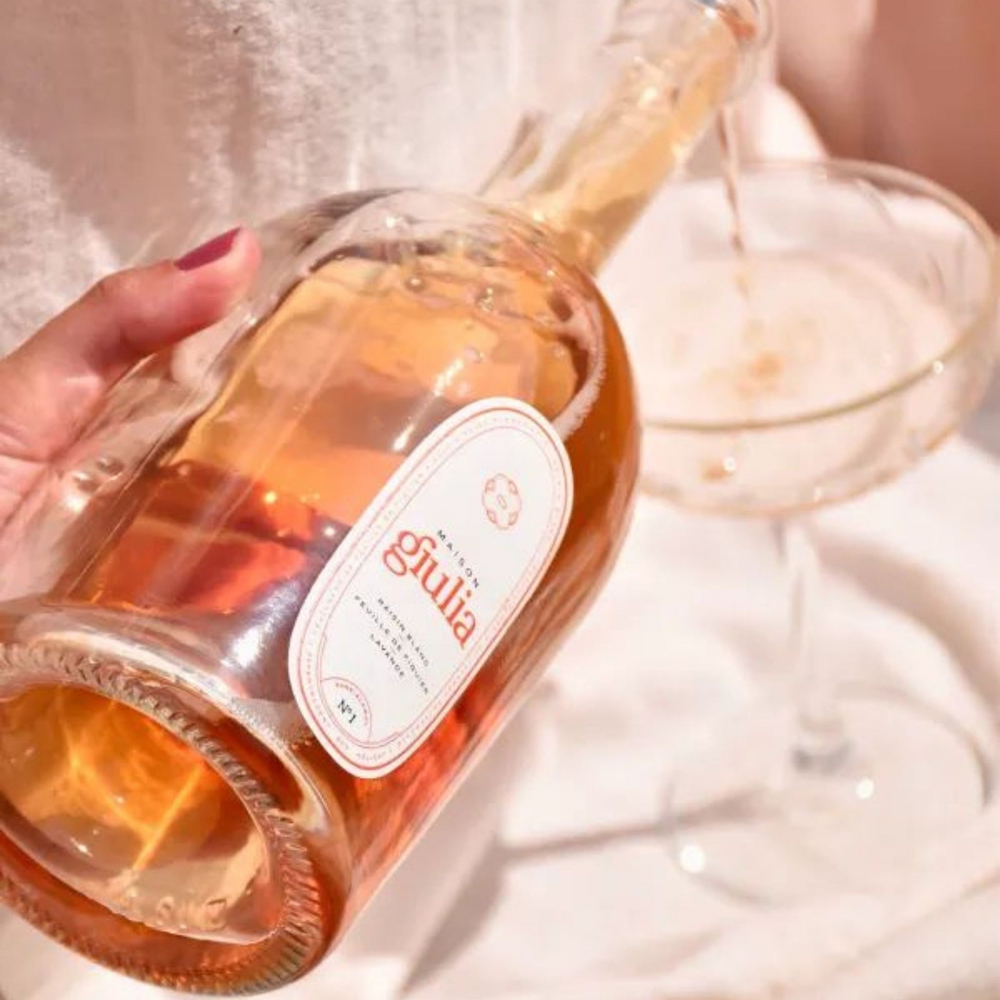 A hand holds a clear glass bottle of Giulia aperitivo, pouring a golden-amber liquid into a coupe glass on a white surface.
