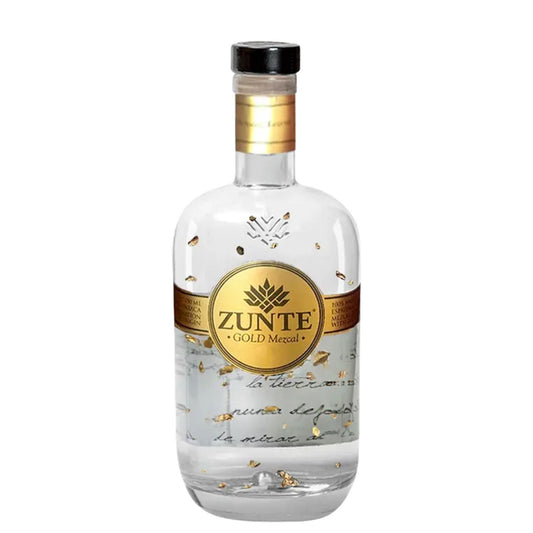 A bottle of Zunte Gold Mezcal on a solid white background. The clear glass bottle features a prominent gold circular label with the brand name 'ZUNTE' and 'GOLD MEZCAL' printed on it, accented with small gold leaf flakes inside.